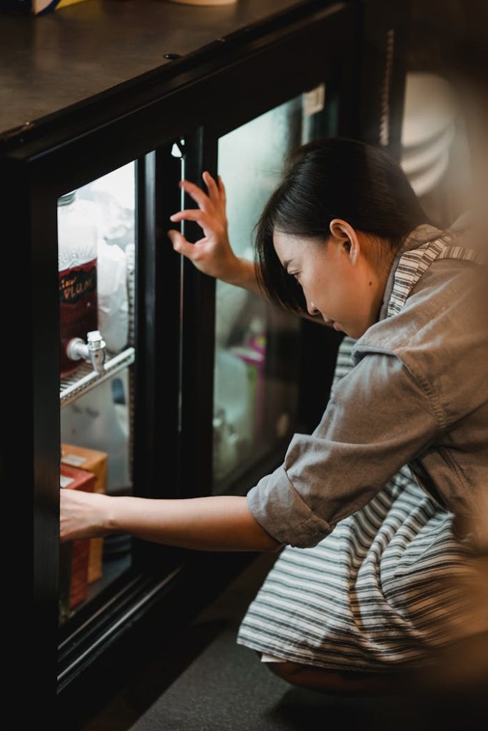 Asian woman crouching to organize items in a modern kitchen pantry.